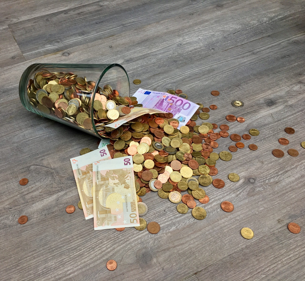 Image of a glass of coins and Euro bills tipped over such that the money lies spilled over a surface